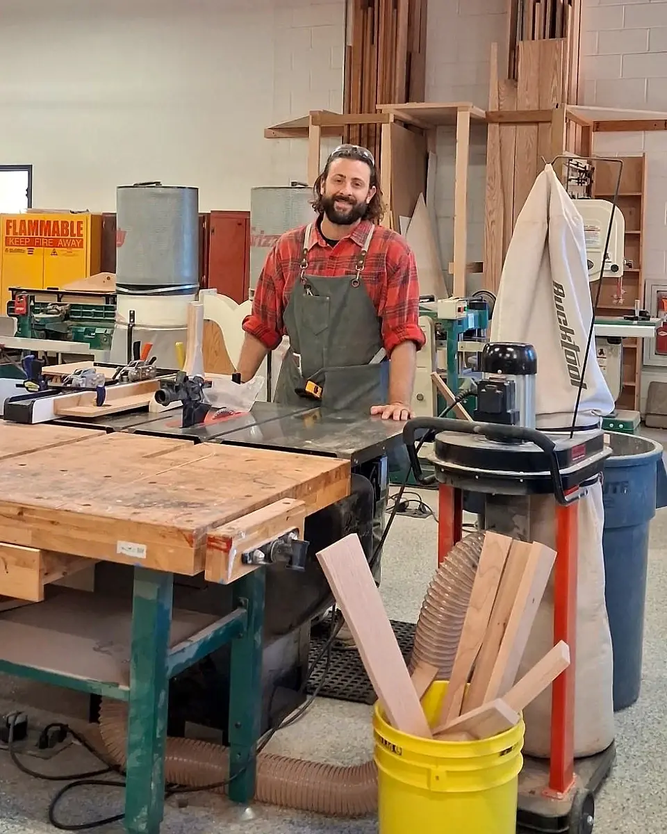 Mr. Spinozzi smiling at a table saw in a busy woodshop, with a workbench, a dust collector and a lumber rack around him